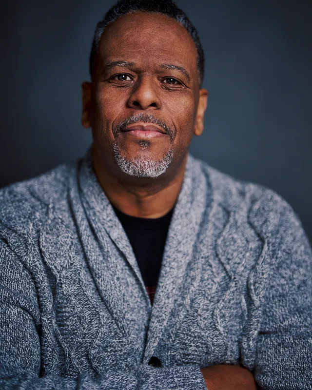 Portrait of C. G. Brown. A Black man with medium brown skin, short salt-and-pepper hair, and a goatee wearing a thick gray knit cardigan sweater over a black t-shirt. His arms are folded and he's looking directly into the camera with a relaxed and open expression.